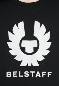 Svart t-shirt med en stor vit logotyp med stiliserade vingar och ett centralt emblem, texten "BELSTAFF" nedanför i fet vit stil.