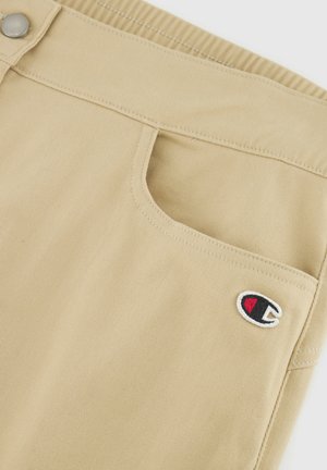 Khakifarbene Shorts aus Denim mit einer Fronttasche und einem gestickten Logo in einem kreisförmigen Design in Schwarz, Rot und Weiß.