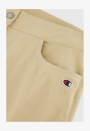 Khakifarbene Shorts aus Denim mit einer Fronttasche und einem gestickten Logo in einem kreisförmigen Design in Schwarz, Rot und Weiß.