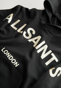 Sudadera negra de tejido suave con texto grande en blanco y dorado metálico que dice "ALLSAINTS" y "LONDRES" en fuentes audaces y contemporáneas.