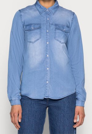 Personne portant une chemise en jean bleu clair boutonnée avec deux poches poitrine et un jean bleu foncé, debout avec les bras relâchés sur les côtés.