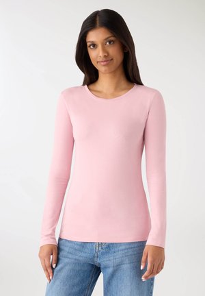 Długi top z długim rękawem w jasnoróżowym materiale, z okrągłym dekoltem i dopasowanym krojem, zestawiony z niebieskimi dżinsami. Gładka tekstura.