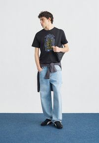 Νεαρός άνδρας που φοράει μαύρο γραφικό μπλουζάκι, ανοιχτό μπλε φαρδιά τζιν, μαύρα αθλητικά παπούτσια, κρατώντας μαύρη τσάντα ώμου, στέκεται σε μπλε χαλί μπροστά σε λευκό τοίχο.