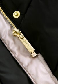 Intérieur d'une veste matelassée noire avec doublure en satin léger. Équipée d'une fermeture éclair plaquée or avec 'Baker' gravé et d'un bouton pression décoratif.