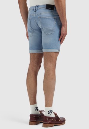 Man wearing light blue rolled-up denim shorts, white socks with dark lettering, and dark brown boat shoes, standing against a plain background.