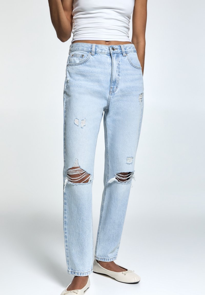 Donna con jeans strappati di un leggero blu e scarpe flat bianche, abbinati a un crop top senza maniche bianco, in piedi contro uno sfondo neutro.