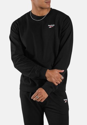 Mann trägt schwarzen Reebok-Sweatshirt und -Hose, hält sein Handgelenk, trägt silberne Kettenhalskette und zwei silberne Ringe vor einem schlichten hellen Hintergrund.