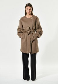Brązowy, wełniany płaszcz z paskiem, okrągłym dekoltem i oversize'owymi rękawami, w połączeniu z czarnymi spodniami i subtelnymi złotymi detalami.