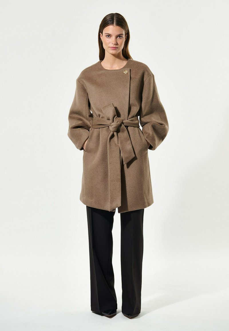 Ruskea vyöllinen villakangastakki, jossa on pyöreä kaula-aukko ja ylisuuret hihat, yhdistettynä mustiin housuihin ja hillityllä kultaisella yksityiskohdalla.