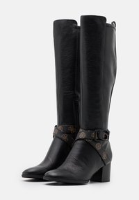 Bottes hautes jusqu'au genou en cuir noir avec un bout légèrement pointu. Elles comportent une sangle à motifs autour de la cheville avec une boucle et une fermeture éclair latérale.