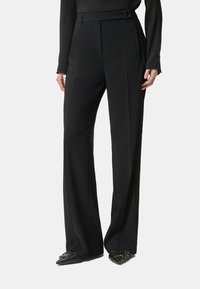 Pantaloni neri su misura con vita alta, gambe larghe e dettaglio cinturino. Tessuto liscio con pieghe a contrasto e tasche laterali.