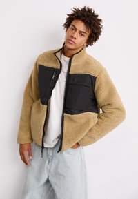 Fleecejacke mit einem Farbblockdesign in Beige und Schwarz, Reißverschluss vorne und einer Brusttasche mit einem Knopfdetail. Weicher Stoff mit einem lockeren Schnitt.