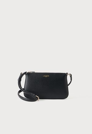 Μαύρη τσάντα Lacoste crossbody με χρυσό λογότυπο, υφή επιφάνειας και ρυθμιζόμενο λουρί σε ανοιχτό φόντο.