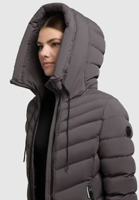 Frau, die eine graue gepolsterte Jacke mit einer übergroßen Kapuze trägt, die zugezippt ist und eine schwarze Innenschicht zeigt, vor einem schlichten hellen Hintergrund.