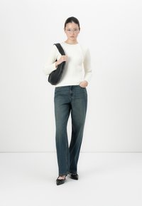 Vrouw in een witte geribbelde trui, blauwe jeans, zwarte puntige hakken, een bril en een zwarte schoudertas, staande tegen een witte achtergrond.