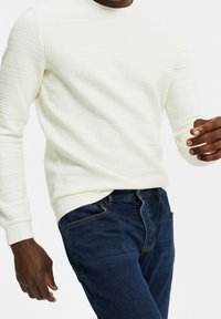 Ljust krämfärgat tröja med texturerat randmönster, rund halsringning och ribbad mudd, i kombination med mörkblå jeans.