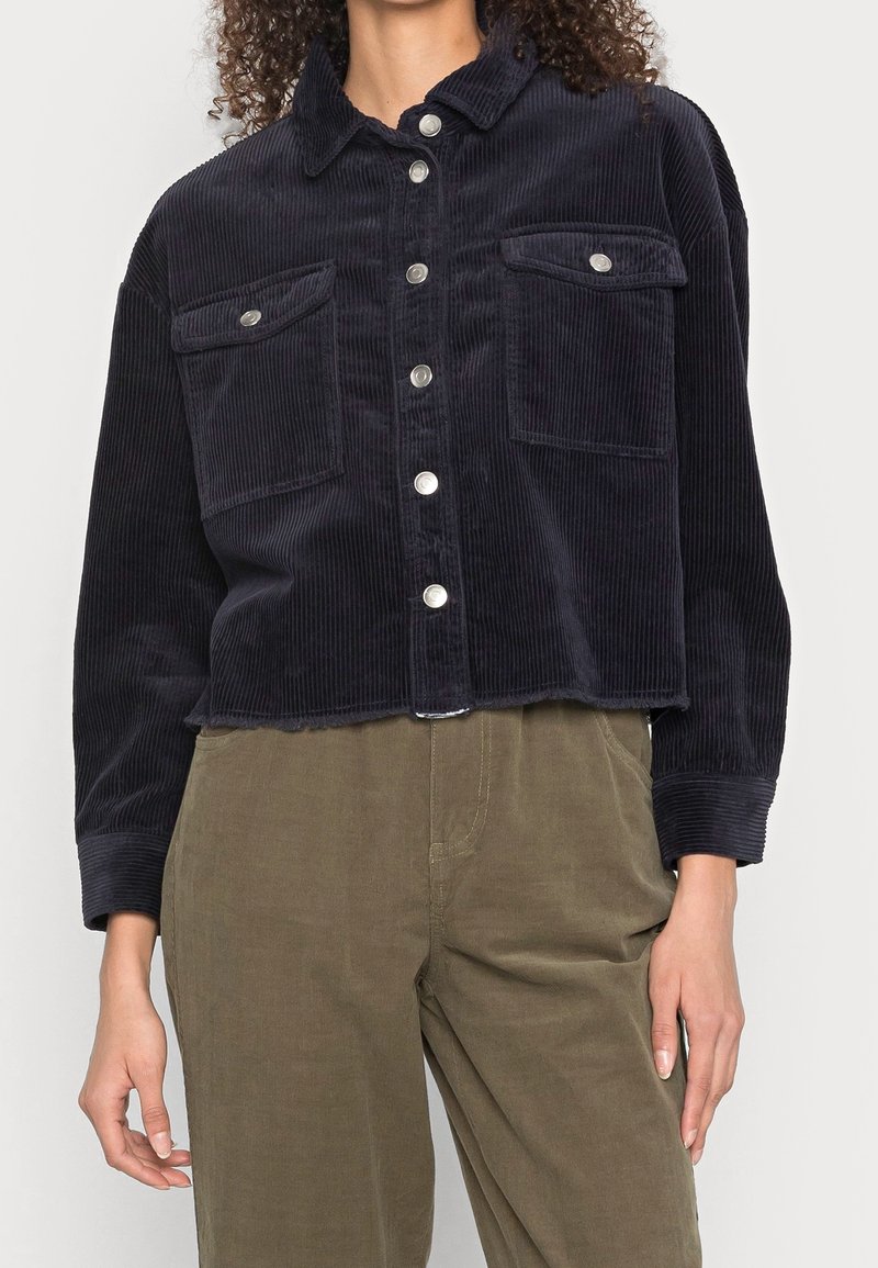 Femme portant une chemise courte noire en velours côtelé à boutons avec poches à rabats sur le devant et un pantalon taille haute vert olive.