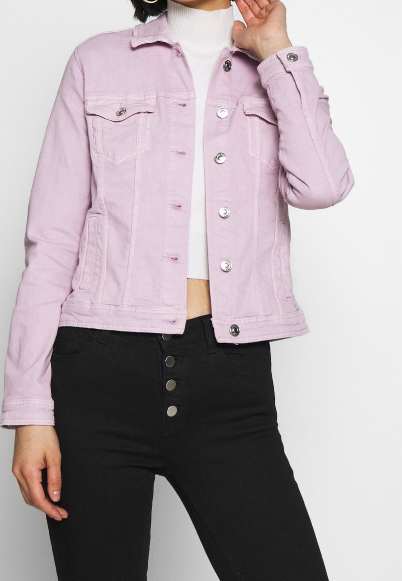 Chaqueta de mezclilla rosa claro con un diseño corto, que presenta bolsillos frontales, botones plateados y una textura ligeramente desgastada. Combinada con pantalones negros.