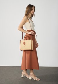 Tan woven handbag with brown leather accents, square shape, and dual handles, worn crossbody. Model in a beige top and rust-colored skirt.