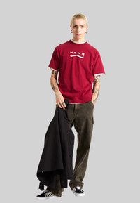 Rød kortærmet T-shirt med hvid Vans-logo, olivengrønne bukser, holder en sort jakke; ternede sort-hvide sneakers.
