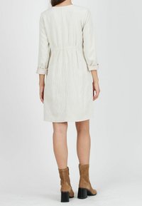 Nainen, joka on pukeutunut vaaleanbeigeen polvipituiseen corduroy-mekkoon, jonka hihat on kääritty ylös, ja hänellä on päällään ruskeat mokka nilkkasaappaat. Nainen seisoo selin yksinkertaista taustaa vasten.