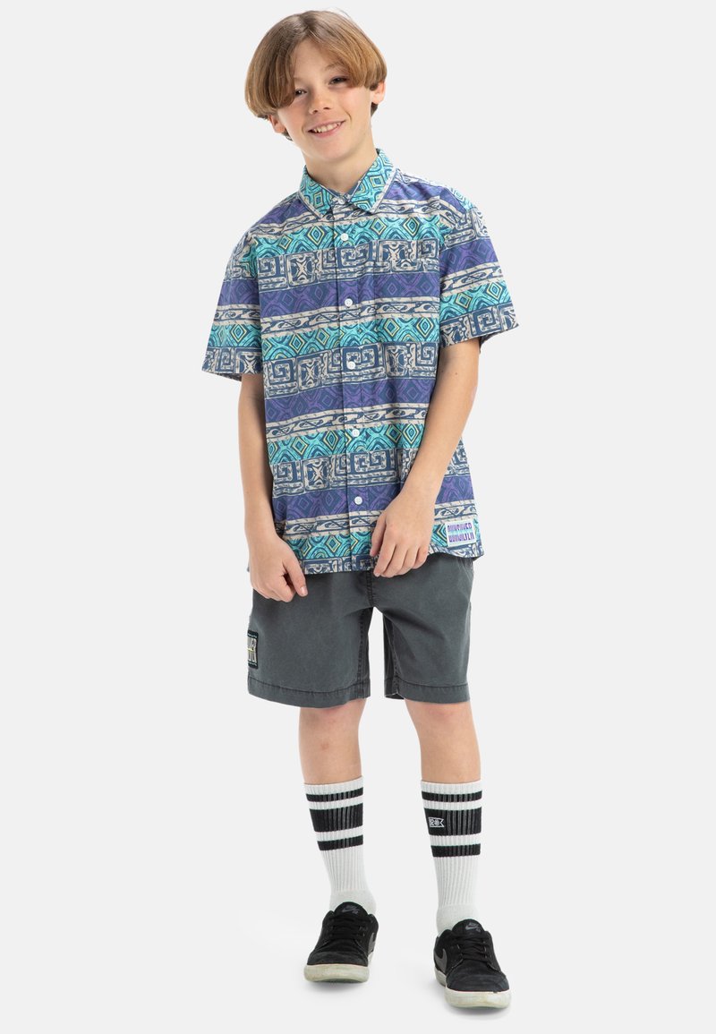Korte mouwen overhemd met blauwe en groene geometrische patronen, gecombineerd met grijze shorts, zwarte sneakers en gestreepte kniekousen.