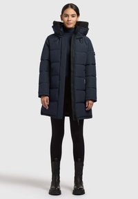 Mørkeblå pufferjakke med fleecefoder i huen, lynlås lukning og sidelommer. Kombineret med sorte leggings og kraftige sorte støvler.