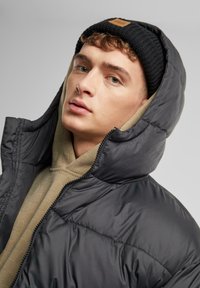Sort puffersjakke med høj krave og lynlås, lagd ovenpå en tan quilted hoodie. Båret med en sort strikket hue.