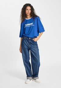 Blå kortärmad T-shirt med vertikal ribbning och vit "hummel PERFORMANCE"-logotyp, kombinerad med löst sittande jeans och ljusa sneakers.