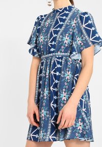 Žena v modrých šatech po kolena s růžovými a tyrkysovými květinovými a geometrickými vzory a krátkými volánkovými rukávy.