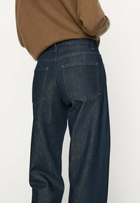 Personne portant un jean taille haute droit bleu foncé et un pull marron, vue de dos sur un fond uni.