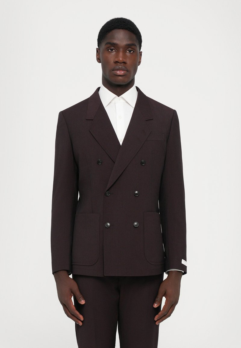 Dunkelbraune, doppelte Blazerjacke mit großen Revers, zwei vorderen Taschen und schwarzen Knöpfen; kombiniert mit einem weißen Hemd.