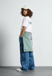 Hvit t-skjorte med grønt logo, grønn hettegenser knyttet rundt livet, løse blå jeans og beige sko. Bakgrunnen er grå.