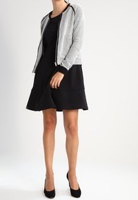 Svart klänning med utsvängd kjol, tillsammans med en grå texturerad zip-up jacka med ett bikakesmönster och svarta detaljer. Svarta högklackade skor.