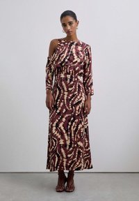 Mujer vestida con un largo vestido estampado en color burdeos con un hombro al descubierto, de pie contra una pared clara y lisa, usando sandalias con pulsera al tobillo.