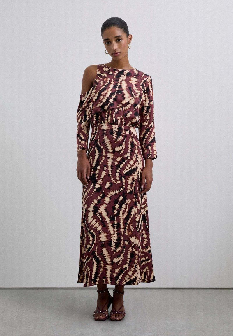 Mujer vestida con un largo vestido estampado en color burdeos con un hombro al descubierto, de pie contra una pared clara y lisa, usando sandalias con pulsera al tobillo.