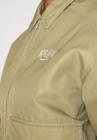 Olive rohelise värvi utilitaarne jakk, millel on luku ees, terav krae ja valge tikandiga logo. Omab taskuid ja nähtavaid õmbluste detaile.