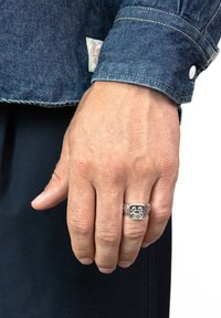 Silverringar med ansiktsdesign, med texturerade accenter, bärs på en hand. Handen är delvis täckt av en mörk denimjacka.