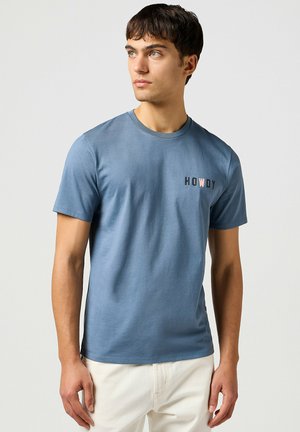 Blauwe katoenen t-shirt met korte mouwen en ronde halslijn. Kenmerkt de tekst "HOWDY" in zwart en roze aan de linkerkant. Soepele textuur.