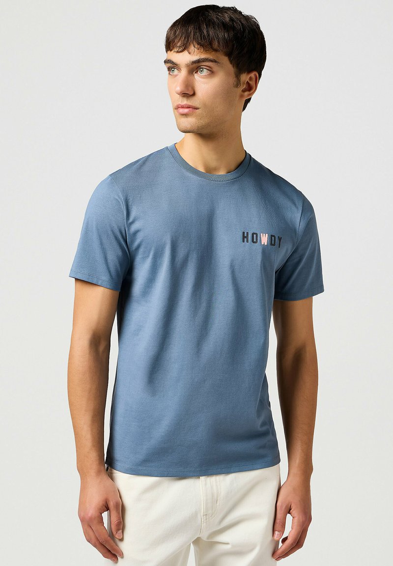 Camiseta de algodón azul con mangas cortas y escote redondo. Presenta el texto "HOWDY" en negro y rosa en el lado izquierdo. Textura suave.
