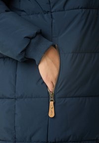 Doudoune bleu marine avec un poignet côtelé, dotée d'une poche zippée avec un tirant en cuir et d'une texture matelassée.