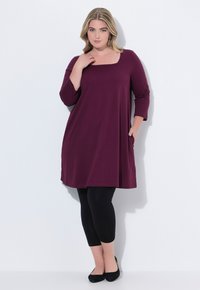 Maroontunika med trekvartærmer, firkantet halsudskæring, sidelommer, parret med sorte leggings og sorte flade sko. Glat tekstur.