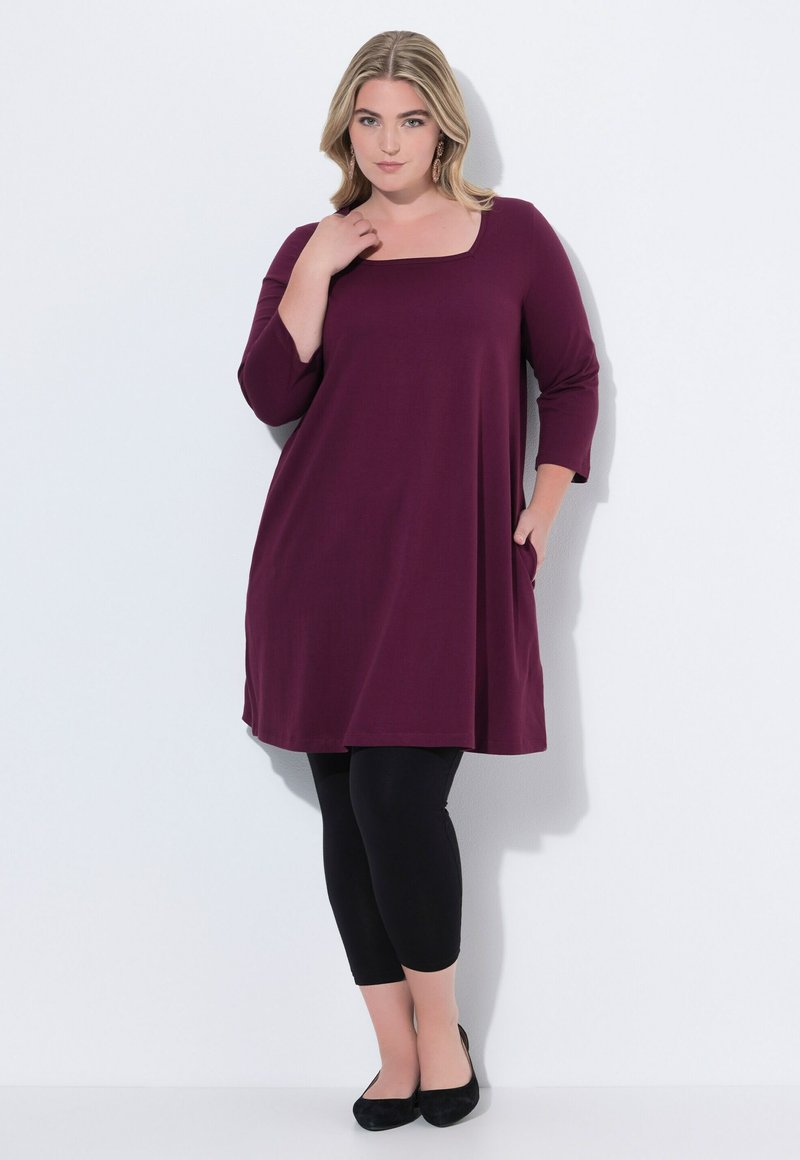 Maroontunika med trekvartærmer, firkantet halsudskæring, sidelommer, parret med sorte leggings og sorte flade sko. Glat tekstur.