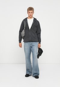 Young man wearing black hoodie, white t-shirt, wide-leg light blue jeans, black shoes, carrying gray shoulder bag and holding black cap.