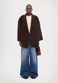 Hnědý vlněný kabát s dvojitým zapínáním, oversized střih, široké modré denimové kalhoty, béžový top, držící hnědou tašku, černé masivní boty.