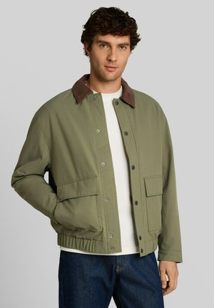 Mann mit lockigem Haar, der eine hellgrüne Jacke mit braunem Kragen, weißem Hemd und blauen Jeans trägt, steht mit einer Hand in der Tasche.