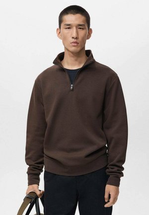 Sudadera marrón con cremallera y cuello alto, hecha de material suave, con mangas largas y un corte relajado. Lleva puesta con pantalones negros y un bolso.