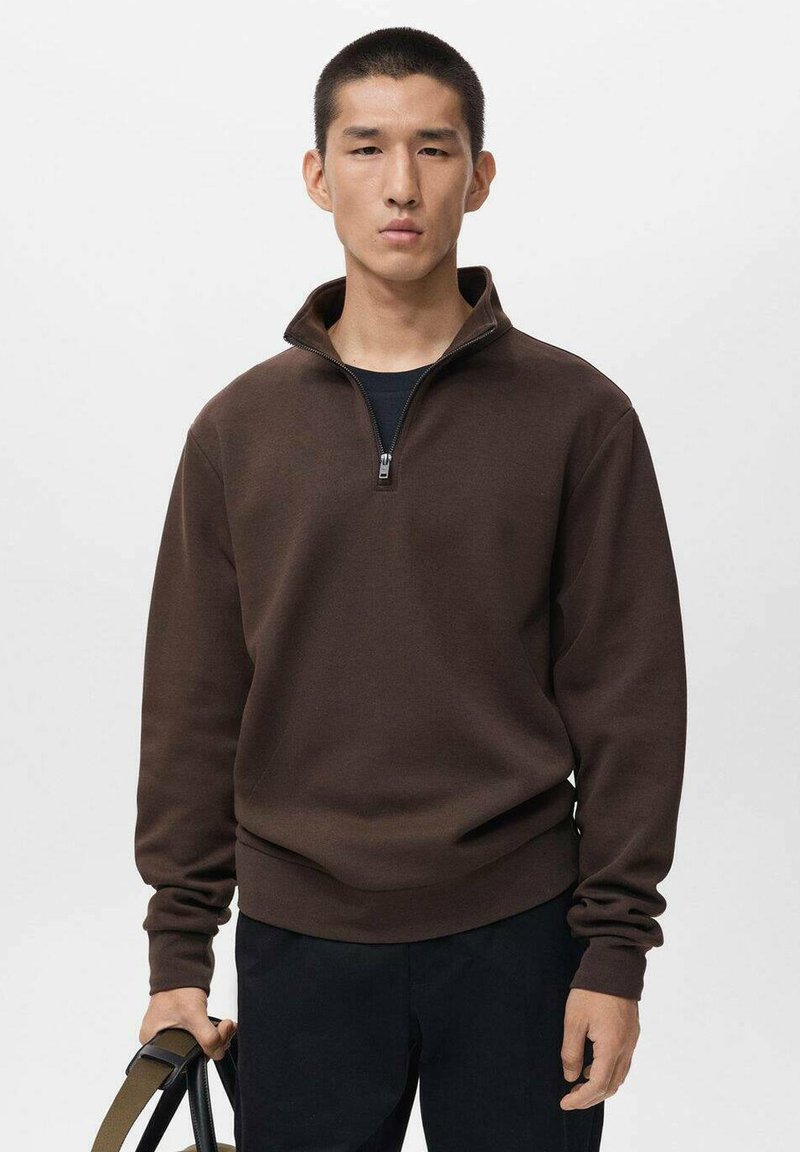 Felpa con zip marrone e colletto alto, realizzata in materiale morbido, con maniche lunghe e vestibilità comoda. Indossata con pantaloni neri e una borsa.