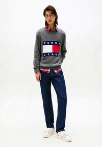 Suéter cinzento de malha com o gráfico vermelho e azul do logo "TOMMY JEANS", sobreposto a uma camisa de xadrez vermelha. Combinado com jeans azuis e sapatilhas brancas.