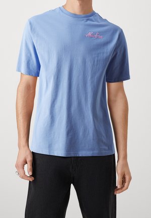 Man wearing a plain light blue short-sleeve t-shirt with pink "New Era" text on chest and black pants against a neutral background.